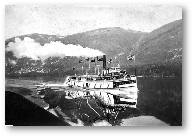 In a spectacular photograph taken from the top deck of the Kokanee, showing the Kaslo cutting the calm water of the West Arm as she gains on her rival. She would go on to win the Victoria Day contest.